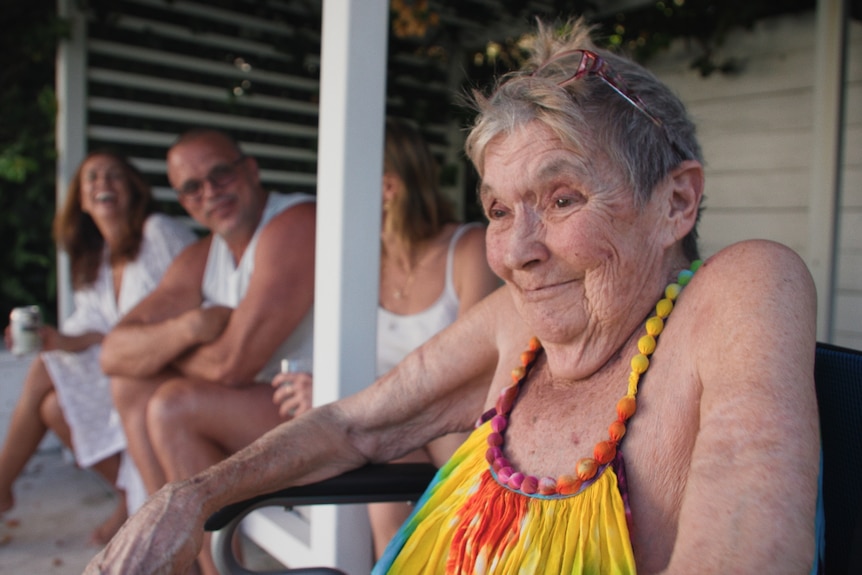 An older woman wearing a rainbow tie-dyed dress sitting in a wheelchair next to her older children who are laughing.