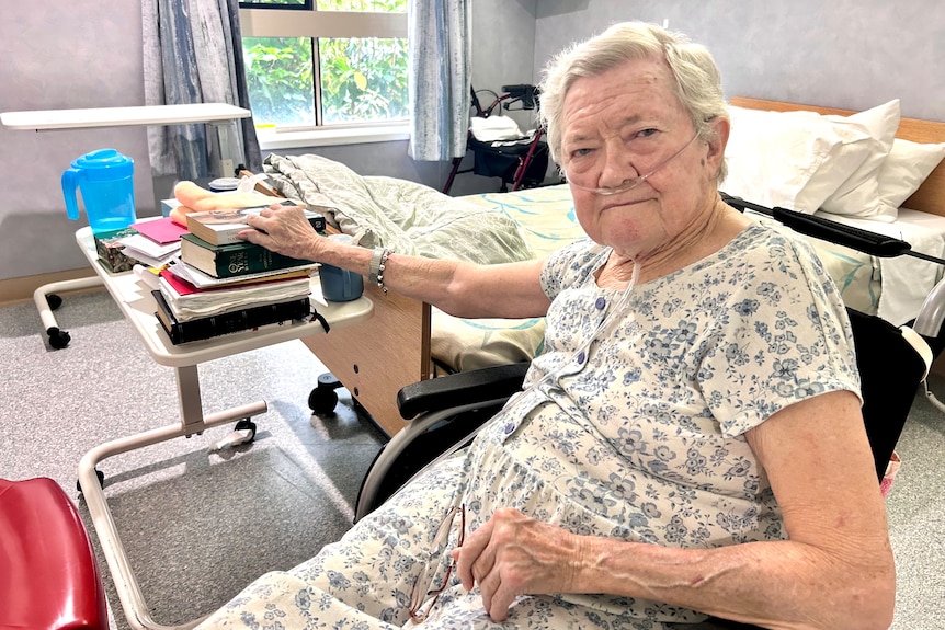 Elderly woman in wheel chair resting arm on table of books in hospital room