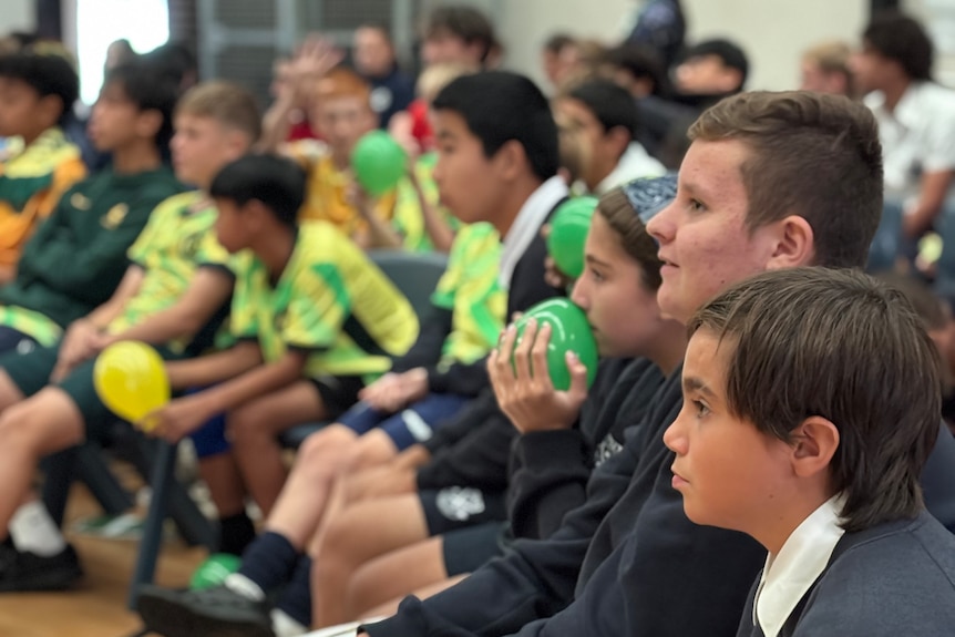 Boys and girls sit on chairs watching the game while holding green and gold balloons looking nervous.