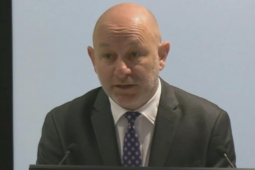 Man in suit speaks into microphone, grey background