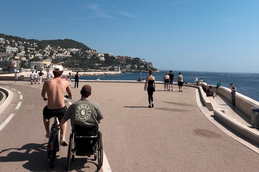 A shot from behind as a man rides a bike with a man in a wheelchair next to him on a path next to the coast.