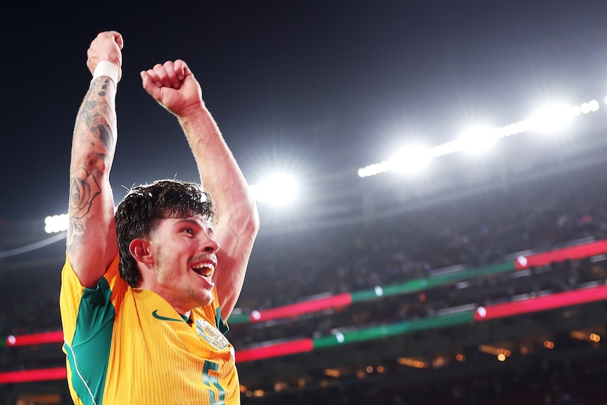 A soccer player in green and gold celebrates a win with arms outstretched