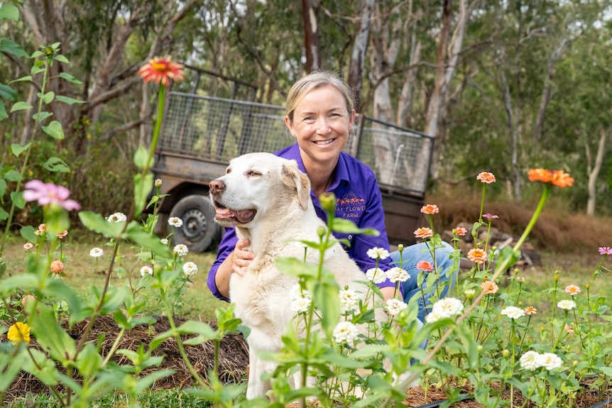 A woman crouches with a dog among flowers.