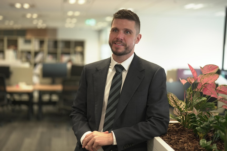 A dark-haired man in a suit stands in an open-plan office.