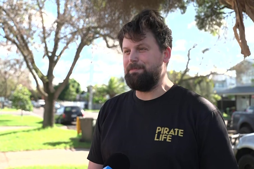 Daniel Livingstone wears a black t-shirt and stands outside his house on the same street as the siege