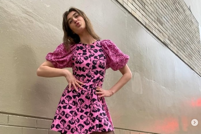 Young woman poses in front of a wall with her hands on her hips, wearing a pink and black patterned dress with puffy sleeves.