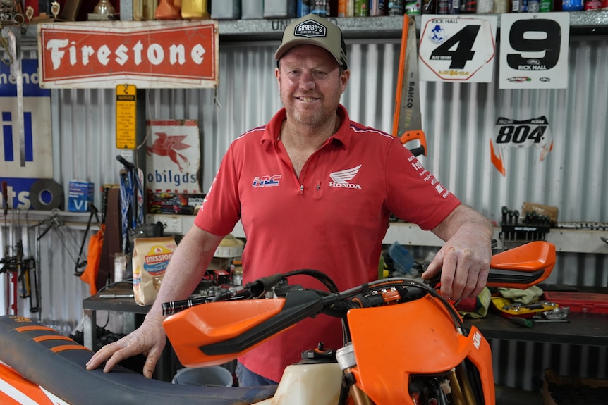A man in a red shirt and cap stands behind an orange motorbike and rests his arm on the handle bars.
