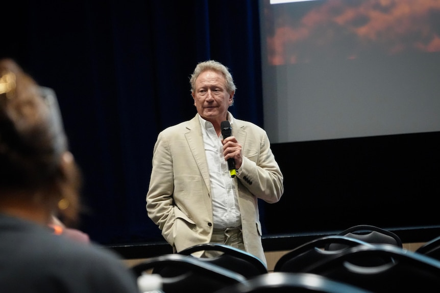 Andrew Forrest speaks at the front of an auditorium with a microphone in his hand and wears a beige suit.