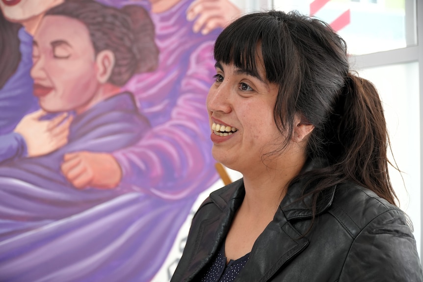A woman with brown hair in a ponytail speaks to someone, with a mural in the background