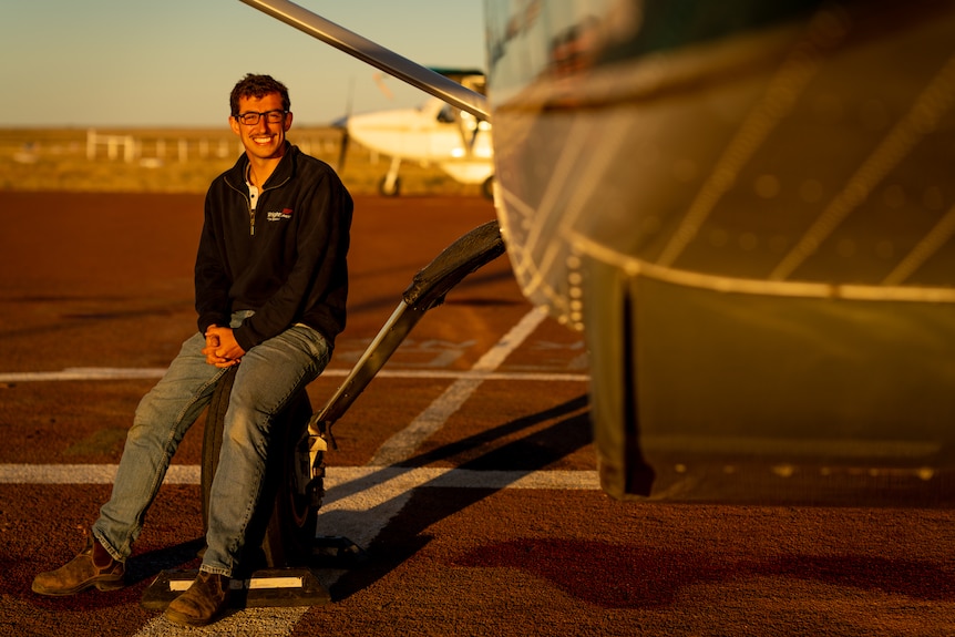 A young man sitting on the tire of a small airplane, smiling at the camera