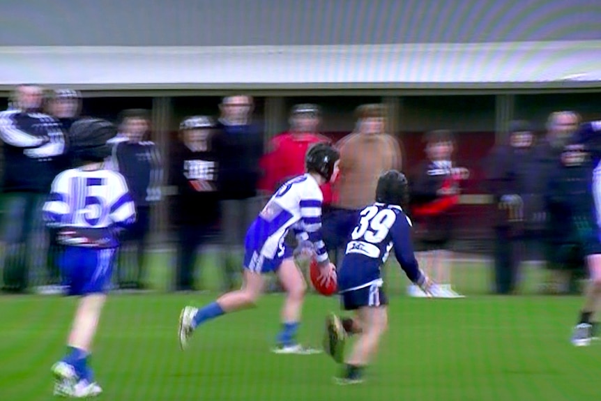 A still from video of boys playing Australian Rules football. One boy is running about to kick the ball.