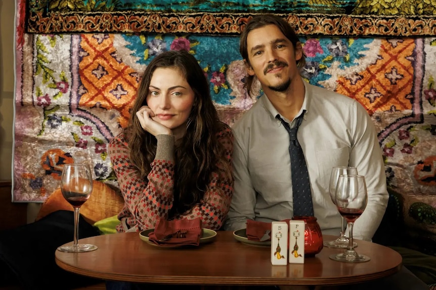 A man and a woman sit at a table and look at the camera.