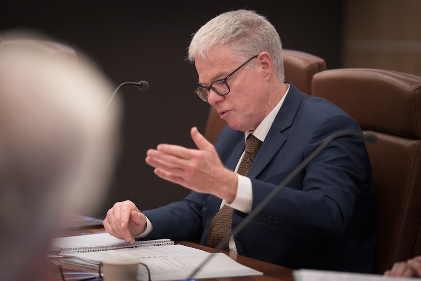 A middle aged man in a dark suit and glasses reads off paper