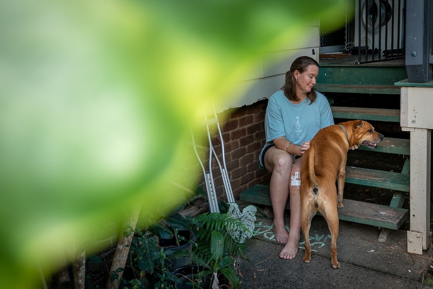 A woman sits on front steps of a wooden house with her dog and crutches leaning against the wall.