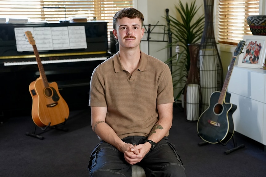 A young man sitting in a room with a piano and two guitars behind him.