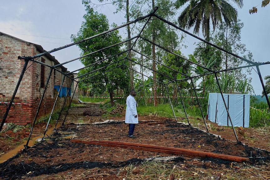 A man in a white lab coat stands inside the metal structure of a tent that has been burned entirely.