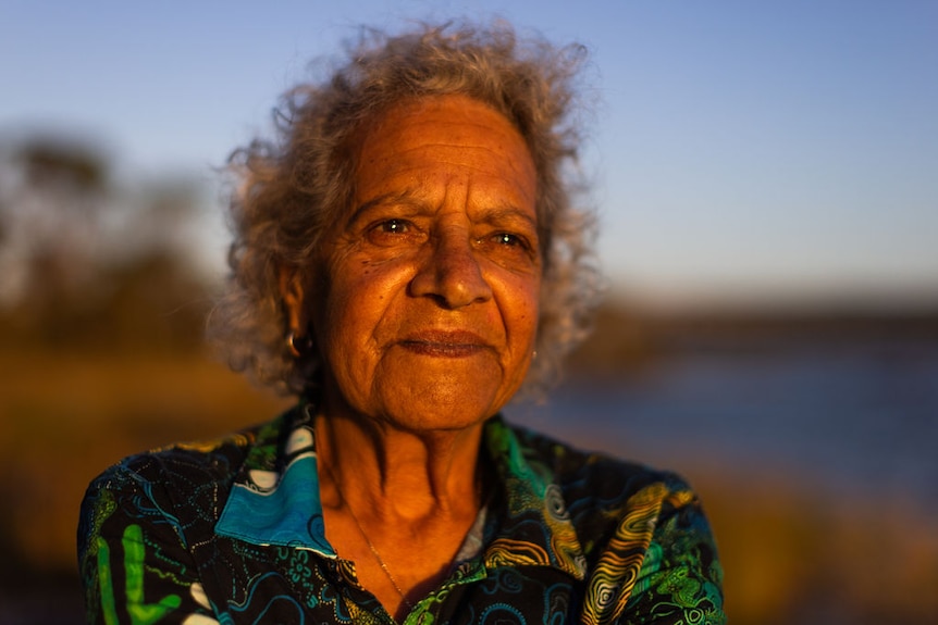 A woman looks into the distance, her face lit up by sun. She wears a blue, green and yellow patterned shirt.