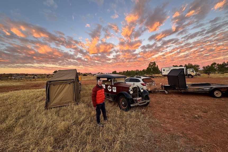 A vintage Studebaker car sits front and centre among a campground in the outback under a beautiful pink setting sky.