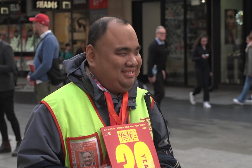 A man with his eyes closed wearing a red lanyard and fluorescent vest holding a red magazine smiles