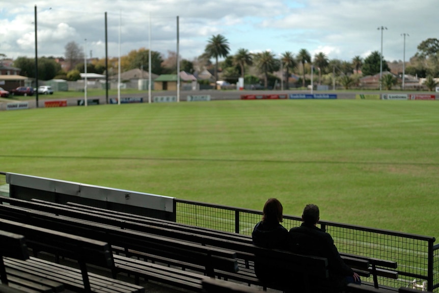 Two people silhouetted sit in the stands of sports field.