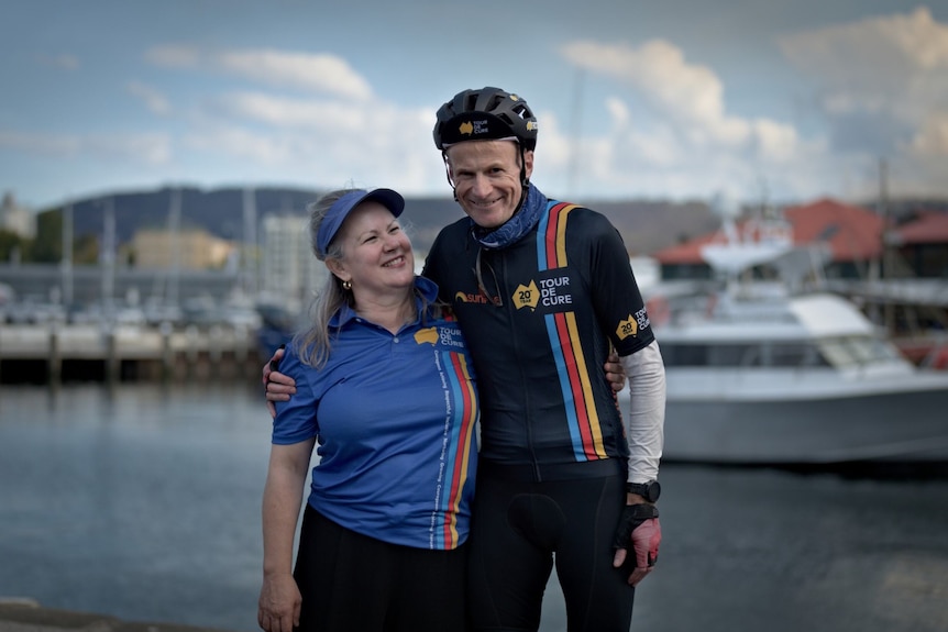 A woman wears a visor hat and blue shirt while looking up at a man with his arm around. He wears cycling gear. They at a marina