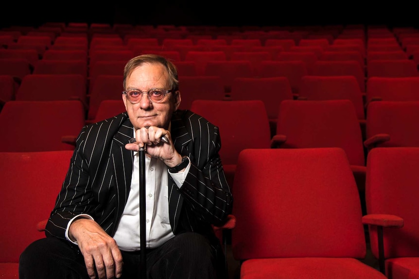 A man wearing glasses and a black striped jacket, sitting in the front row of a theatre with red seats.
