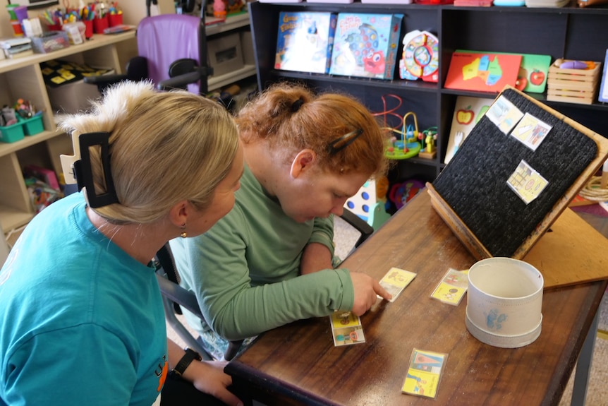 A young woman points to a card on her desk with a board they put cards onto and support worker sitting with her