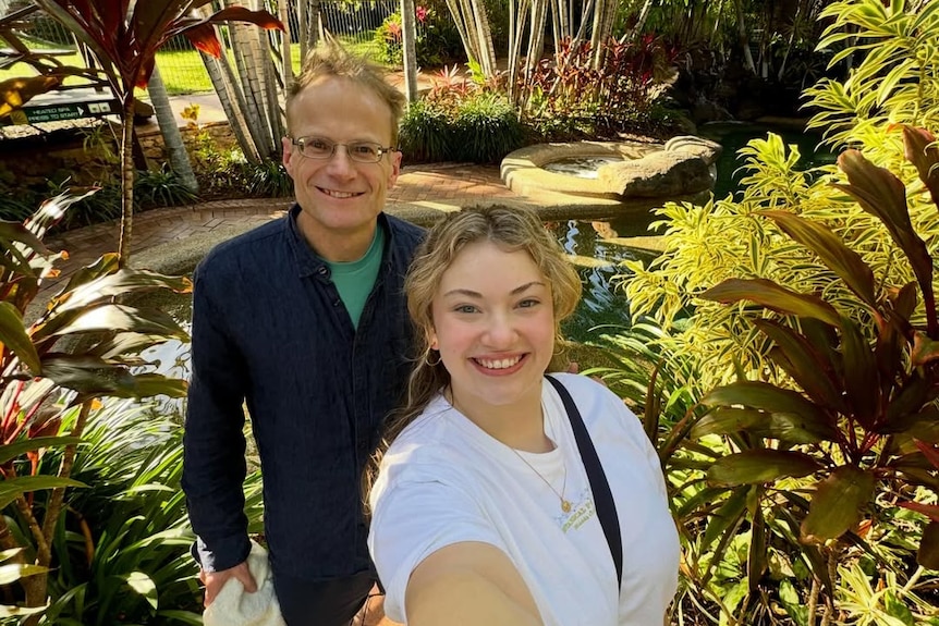 Young woman takes a selfie photo with her father standing behind smiling. They stand with trees and plants behind them