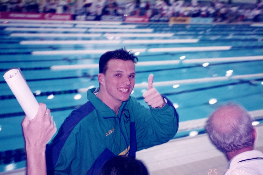 A young Scott Miller wearing an Australian green and gold tracksuit giving the thumbs up in front of a swimming pool.