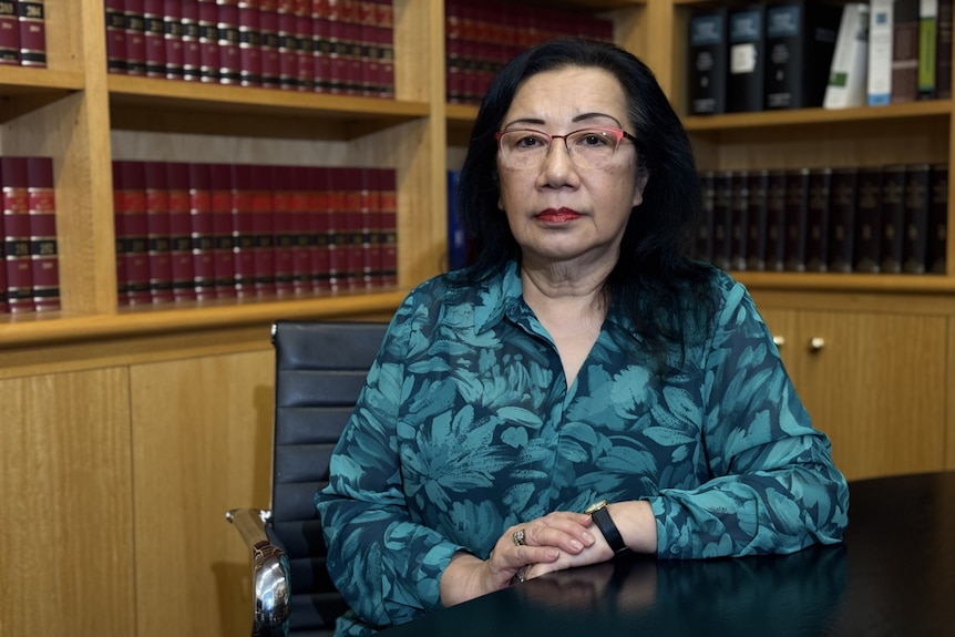 A profile shot of a woman in a blue flower-patterned shirt, glasses and black hair, seated at a table with rows of books behi