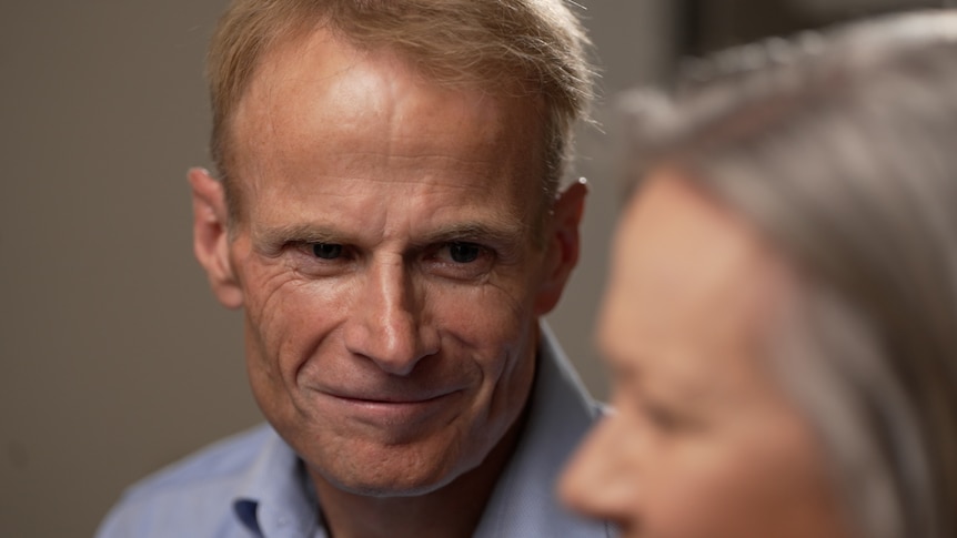 A man with wispy strawberry blonde, short hair looks at a woman who is blurred in foreground