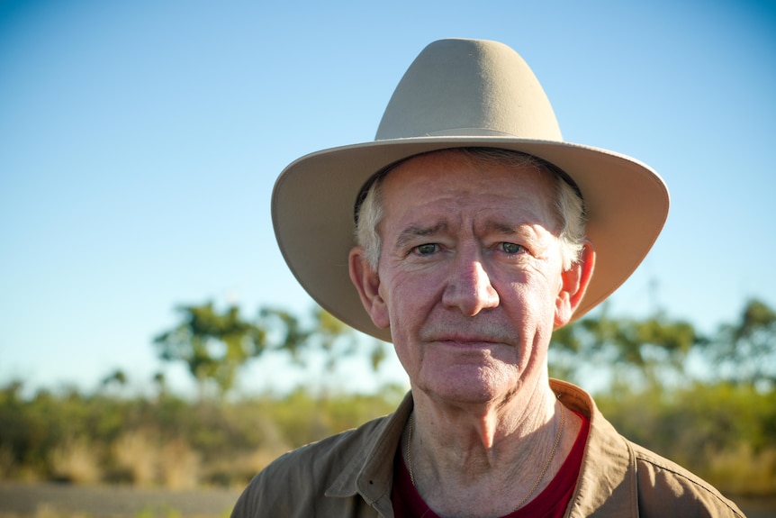 A older man with white hair standing in front of shrubs with a cowboy hat on