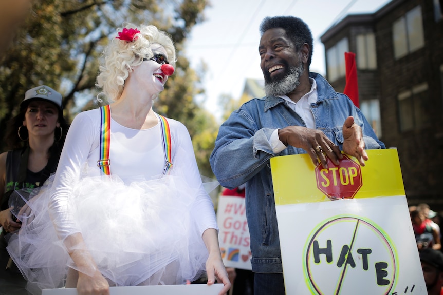 A woman dressed as a clown laughs next to a man holding a stop hate sign.