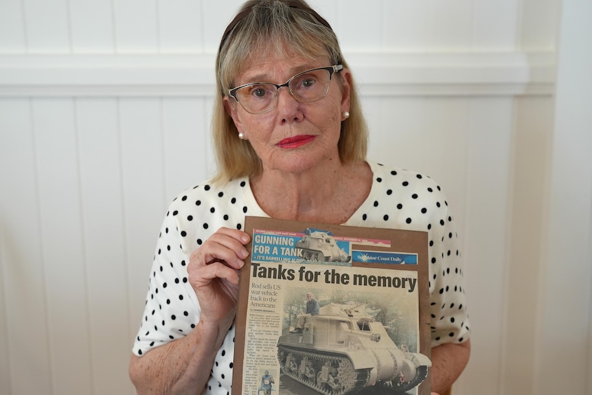A woman with glasses looking sad, holding a newspaper image of a man in a military tank.