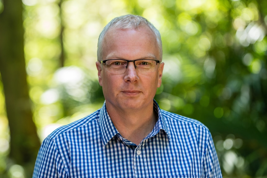 A man with short, grey hair, wearing a blue checked shirt, standing outside in front of trees.