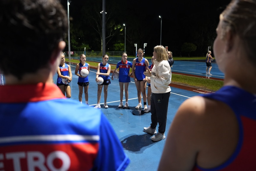 Stacey Marinkovich talks to young netball players who are standing around in a huddle listening on the netball courts.