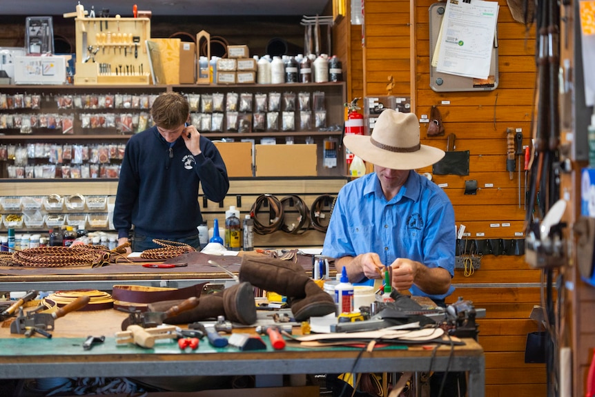 a man with a light blue shirt and wide brim hat inside in a pine studio working on leather