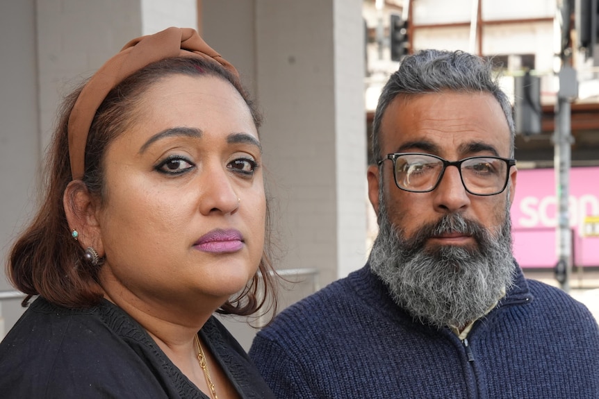 A dark-haired woman and a bearded man, both looking sad, near a street corner in a city.