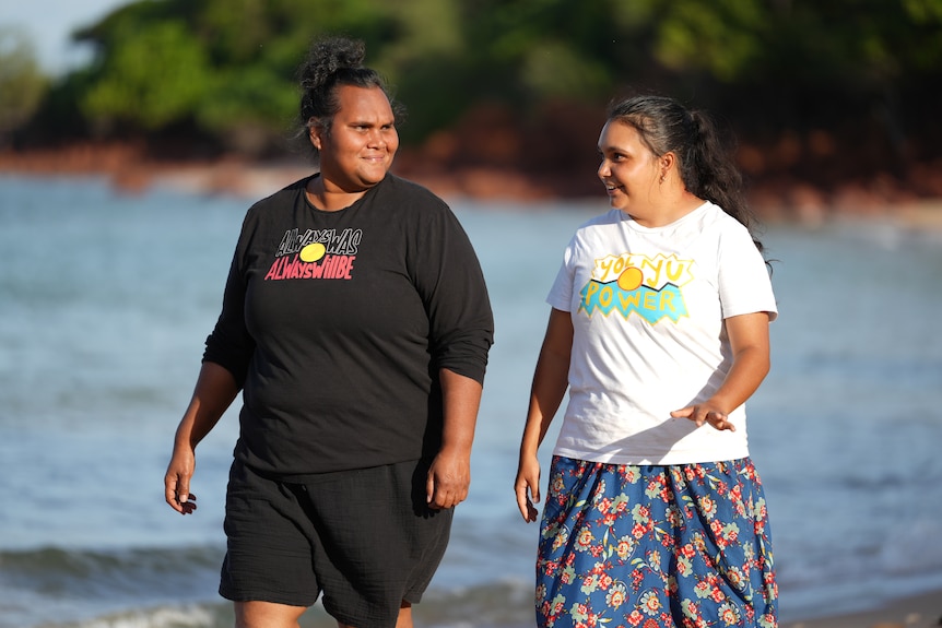 Two Indigenous women walking along the beach, they are chatting to each other and smiling.
