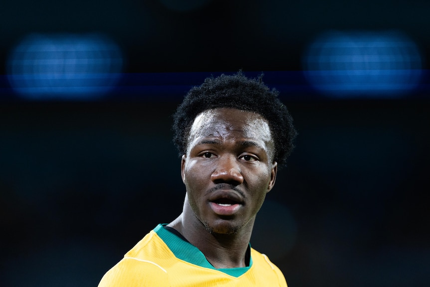 Nestory Irankunda of the Socceroos wears yellow jersey, background dark with blue light.