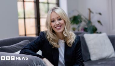 Emma Kay is seen sitting on a grey sofa with white cushions. She has long, wavy blonde hair and is wearing a navy suit jacket and a white t-shirt.