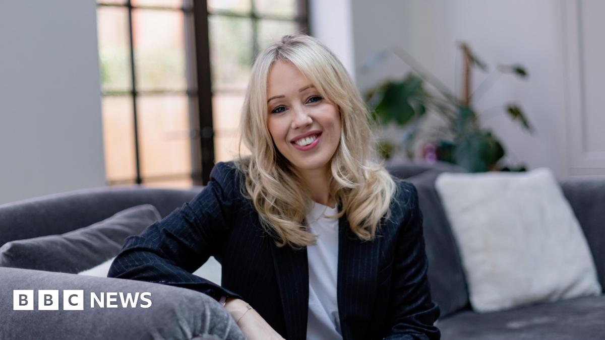 Emma Kay is seen sitting on a grey sofa with white cushions. She has long, wavy blonde hair and is wearing a navy suit jacket and a white t-shirt.