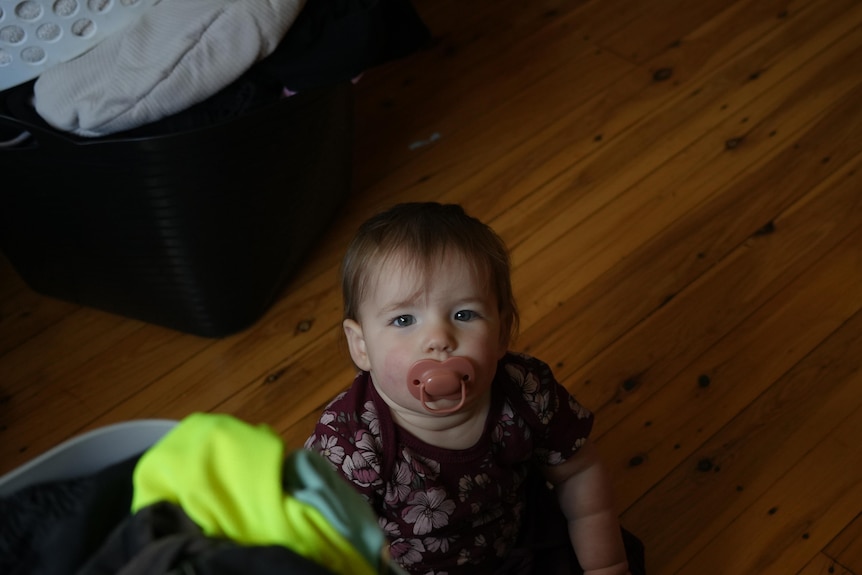 A toddler sucks a dummy while sitting on the floor looking up at the camera.