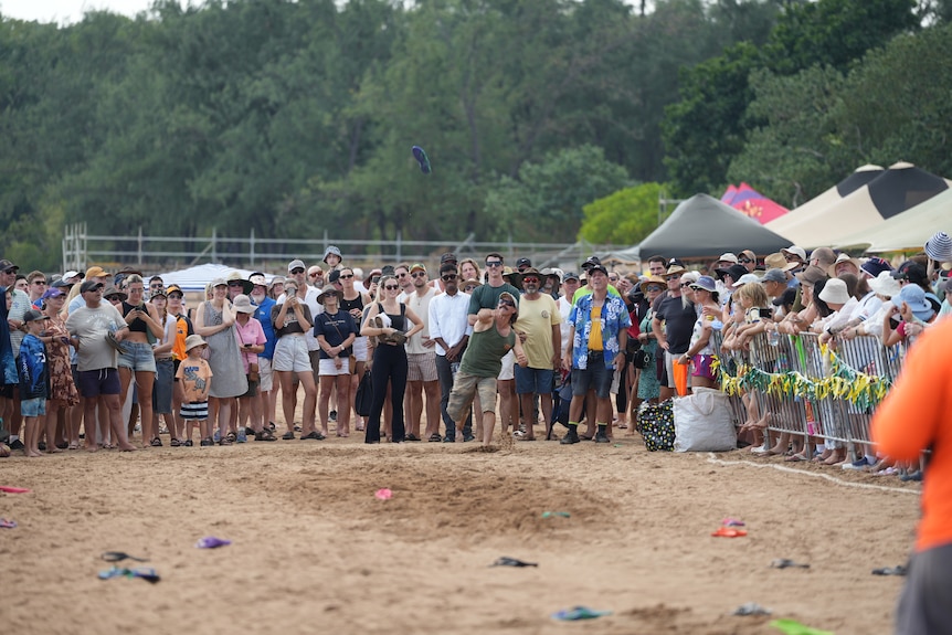 A man throws a thong at a thong-throwing contest in front of a large crowd on a beach