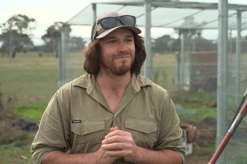 A man with brown hair wearing a cap stands in front of outdoor wildlife pens.