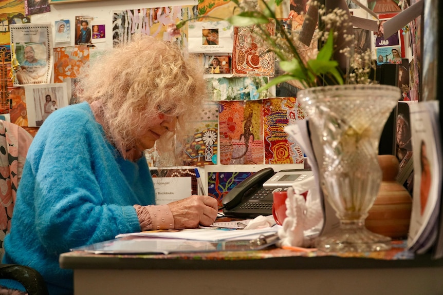 Dr Buchanan sits at a table in a light blue sweater writing.