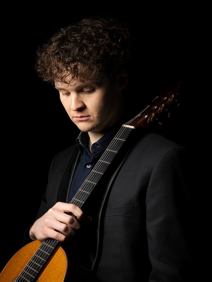 Studio portrait of curly-haired classical guitarist, Connor Whyte holding instrument against shoulder, on a black background.