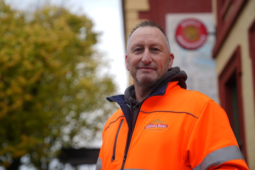 A middle-aged man in orange hi-vis stands in front of a red-brick building with a Boags sign