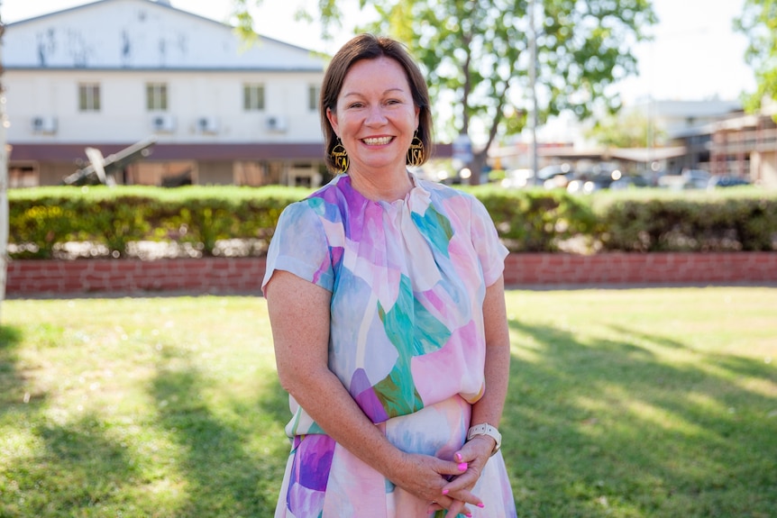 A woman wears a colourful top with green pink and purple in front of a garden. She is smiling.