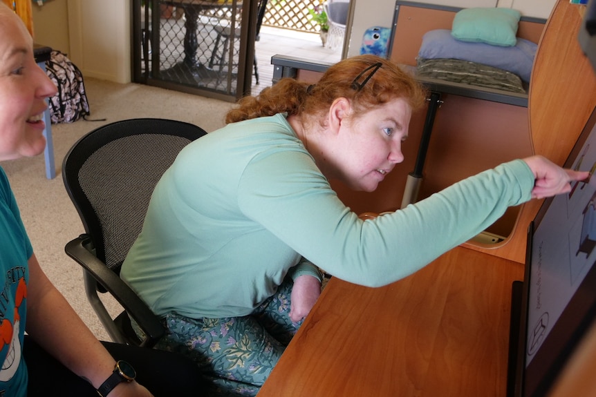 A woman with a disability sitting at her computer pointing to the screen with her support worker smiling beside her